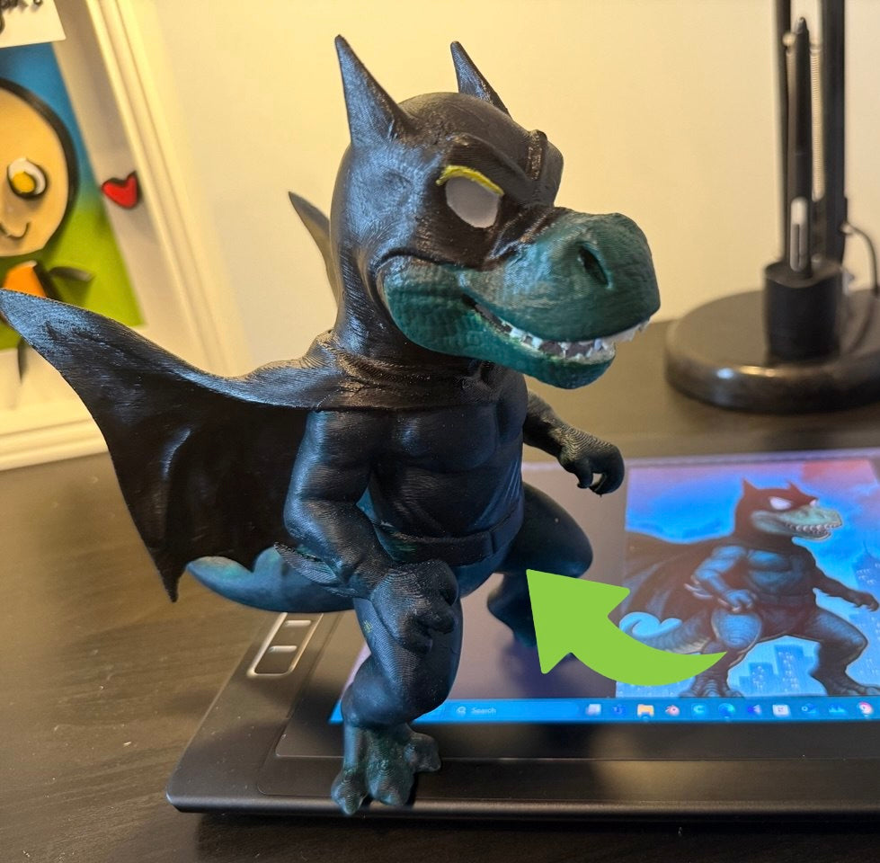 The image shows a 3D-printed toy resembling a batman figure with a dragon-like tail and wings. The toy is standing on a surface in front of a laptop displaying a colorful image.