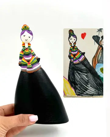 A hand holds a figurine of a woman with a colorful striped top and a long black skirt, recreated from a child's drawing shown beside it. The figurine has a simple face, a bun hairstyle, and a small crown.
