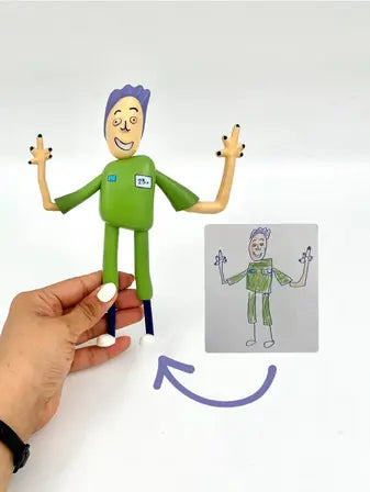 A hand holds a colorful figurine modeled after a child's drawing, which is shown beside it. The figurine has purple hair, a green outfit, and is smiling with both arms raised, mimicking the drawing.