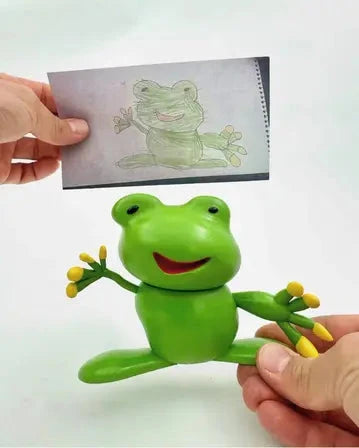 A person holds a green frog figurine with a big smile and yellow-tipped fingers, which closely resembles a child's drawing of a frog shown above it.