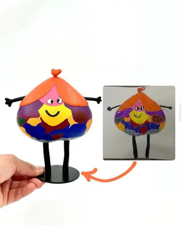 A hand holds a colorful, heart-shaped figurine with a smiling face, modeled after a child's drawing shown beside it. The figurine has a red heart on top, orange and purple hair, and a multicolored body with hearts.