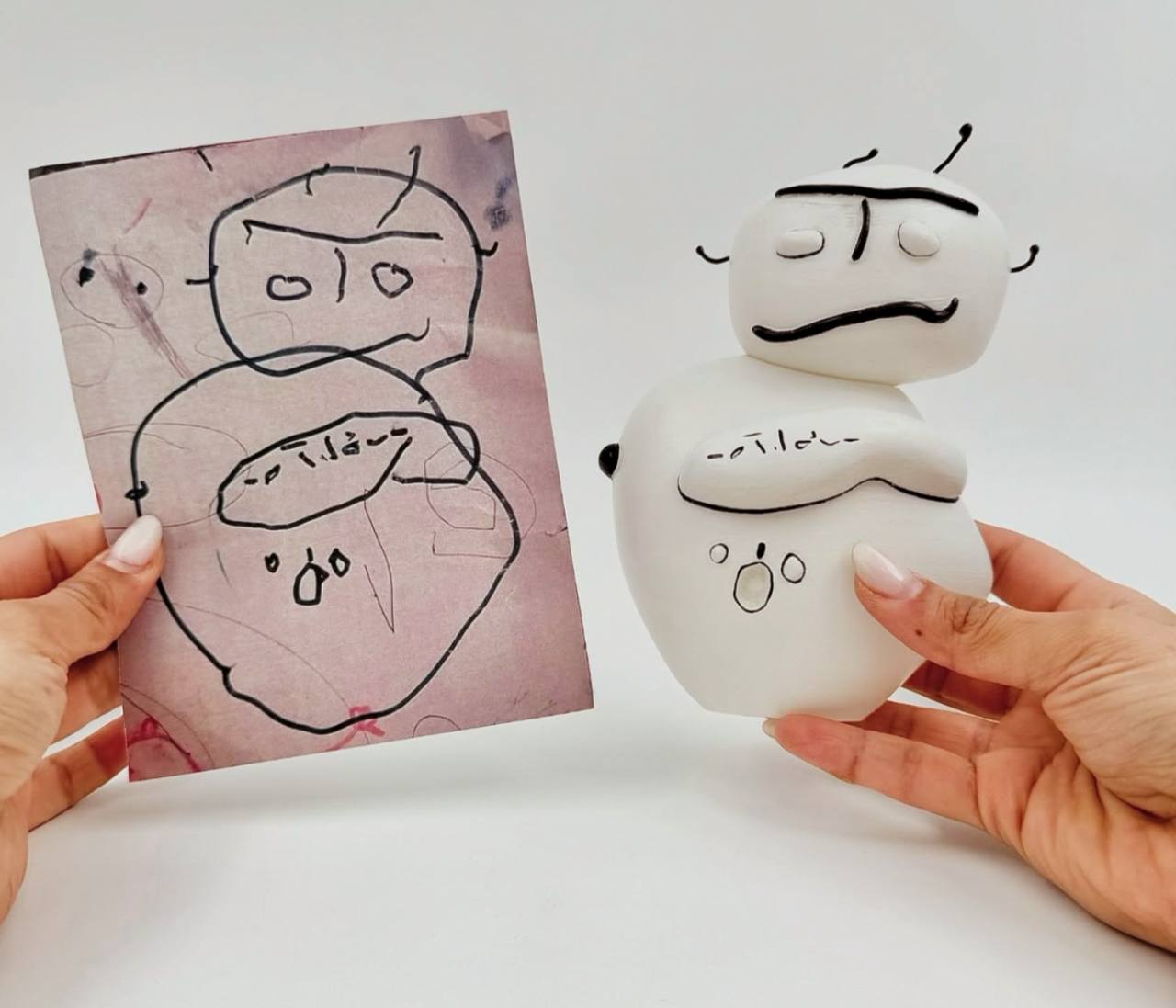 The image shows a hand holding a drawing of a robot with two heads and antennae, and another hand holding a 3D printed version of the same robot.